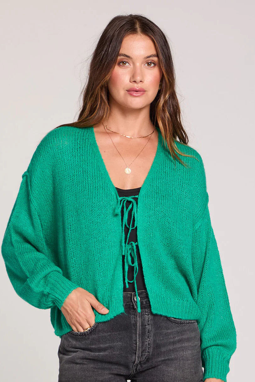 Woman wearing a green cardigan over a black top and denim shorts on a plain background