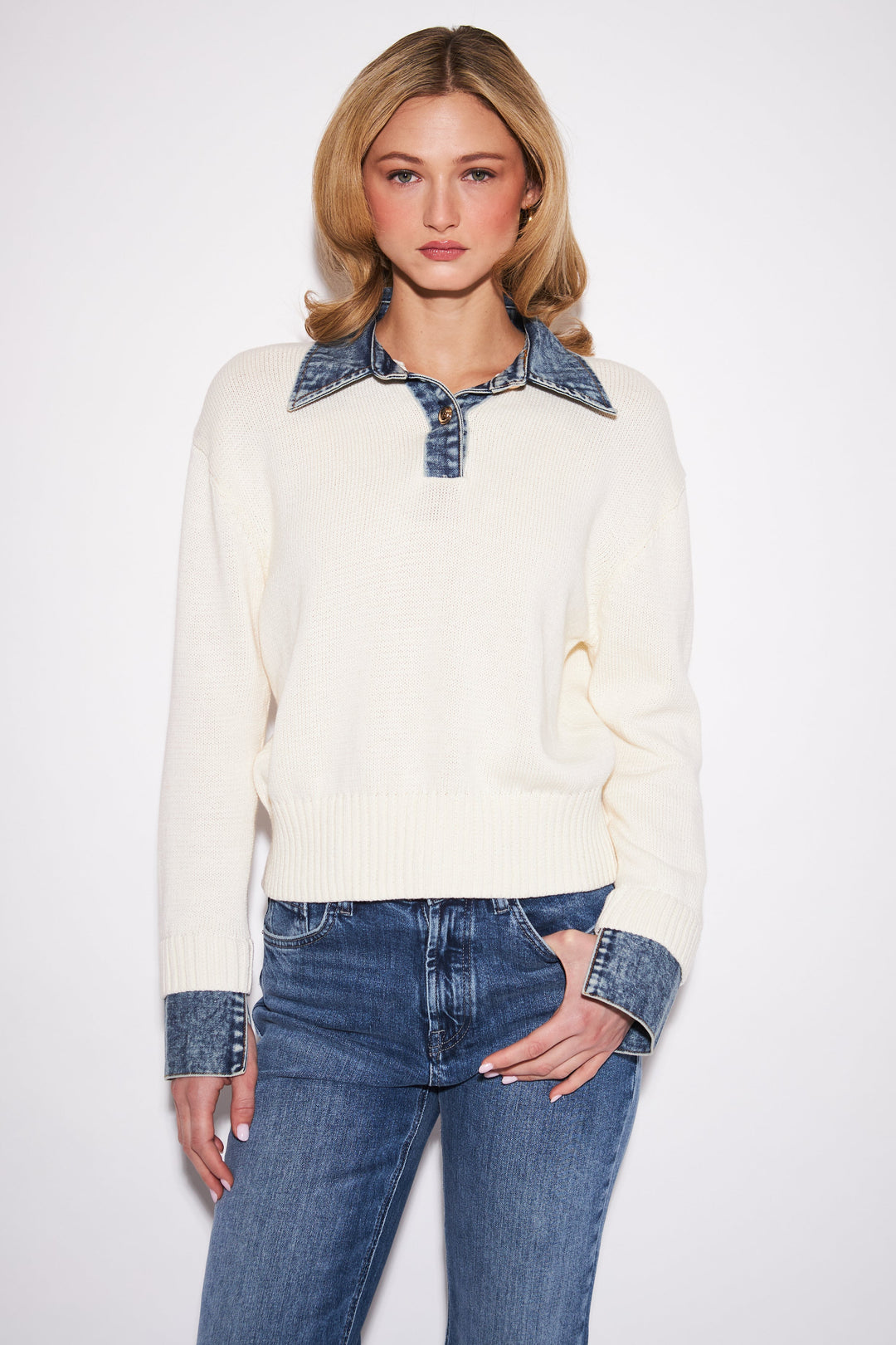 A woman modeling a cream knit sweater with contrasting blue denim collar and cuff details, paired with blue jeans, against a plain light gray studio background.