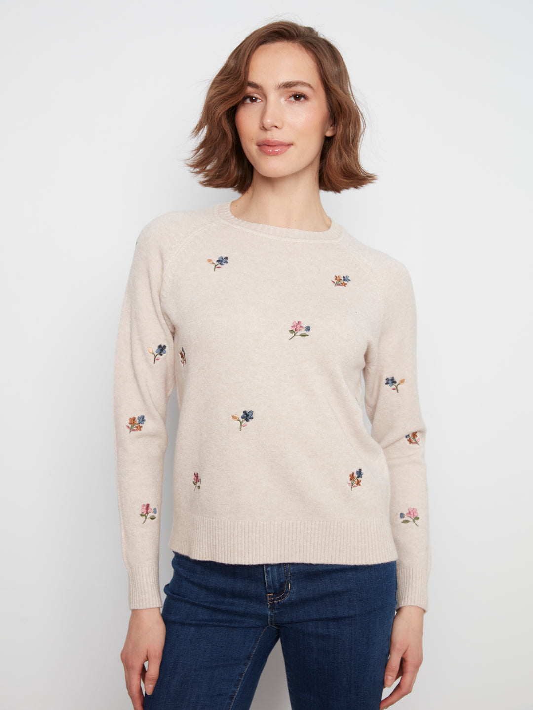 A woman wearing a cream-colored crewneck sweater with small embroidered floral motifs, paired with dark blue jeans, against a plain light gray background.