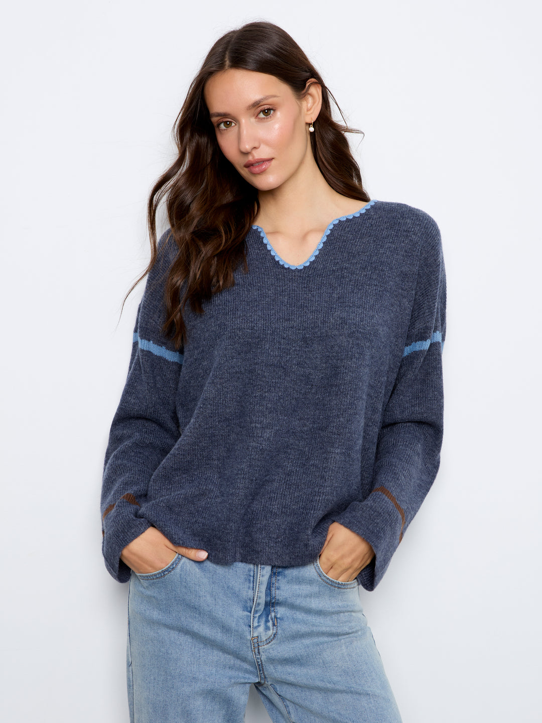 A woman wearing a navy blue knit sweater with light blue trim details on the V-neck and sleeves, paired with light-wash blue jeans, against a plain white studio background.