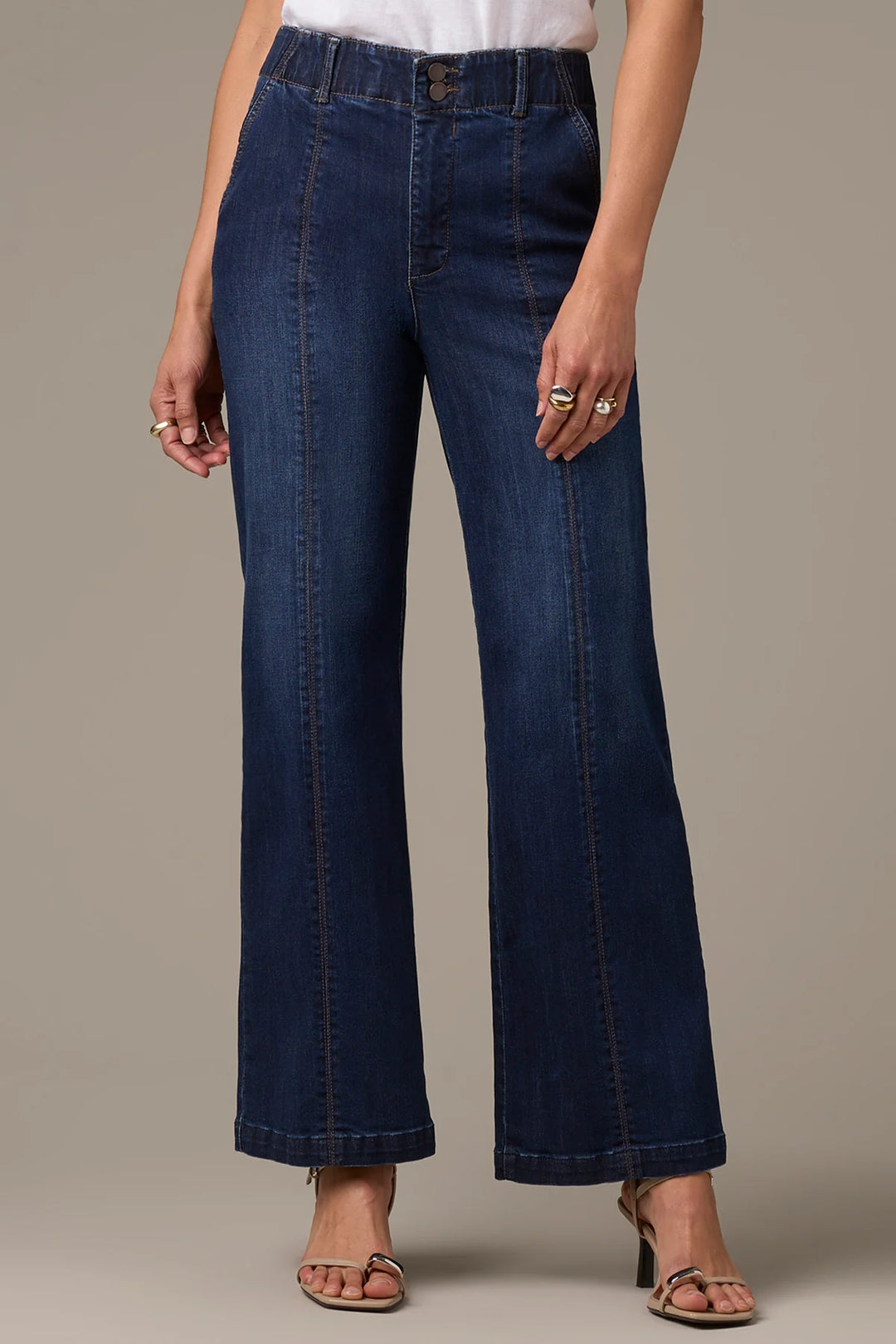 A model wearing a pair of dark blue wide-leg denim jeans, styled with a white top and metallic heeled sandals against a solid beige studio background.