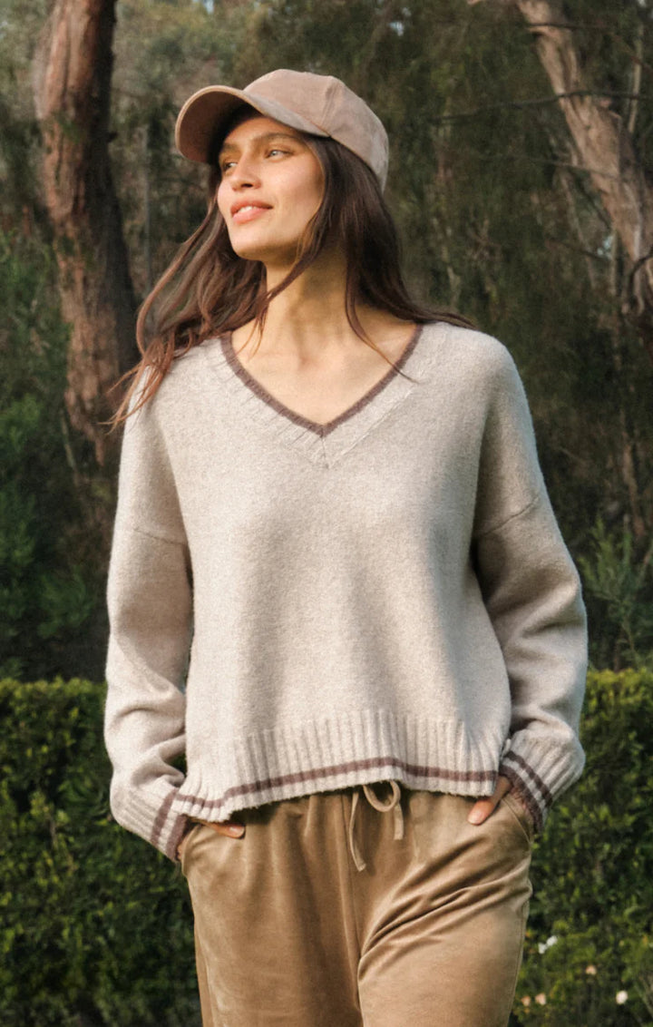A woman wearing a beige v-neck knit sweater with brown trim, tan pants, and a matching beige cap, standing outdoors in front of trees and hedges.