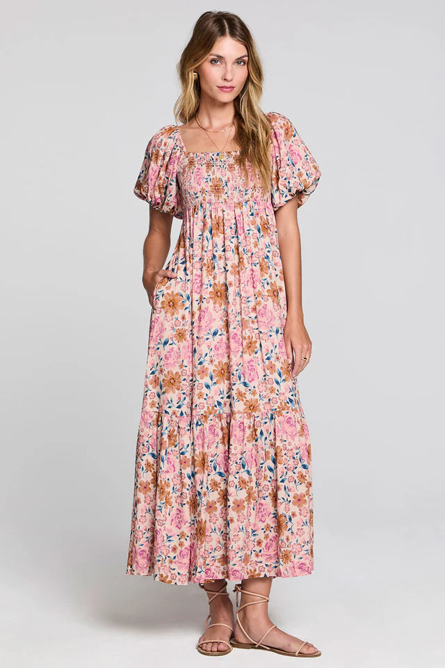 A woman modeling a long-sleeved maxi dress with a pink, orange, and blue floral print, featuring puff sleeves and a square neckline, set against a plain light gray studio background.