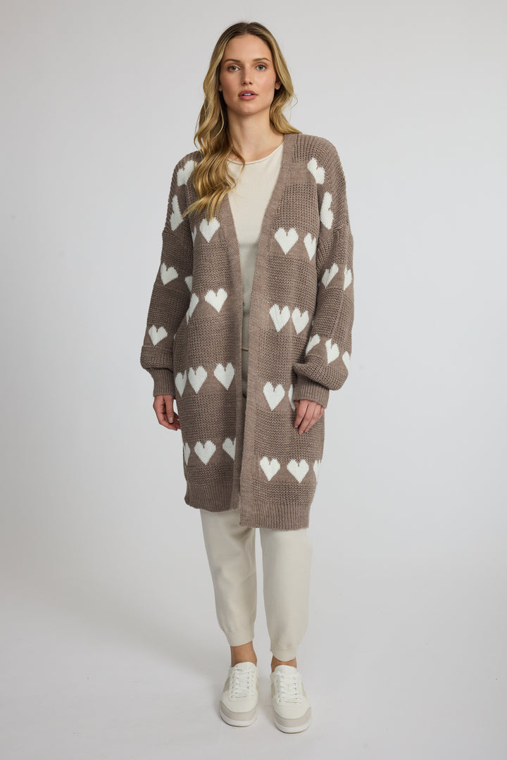 A woman stands against a light gray studio background wearing a long taupe knit cardigan with a white heart pattern, a cream top, cream pants, and white sneakers.