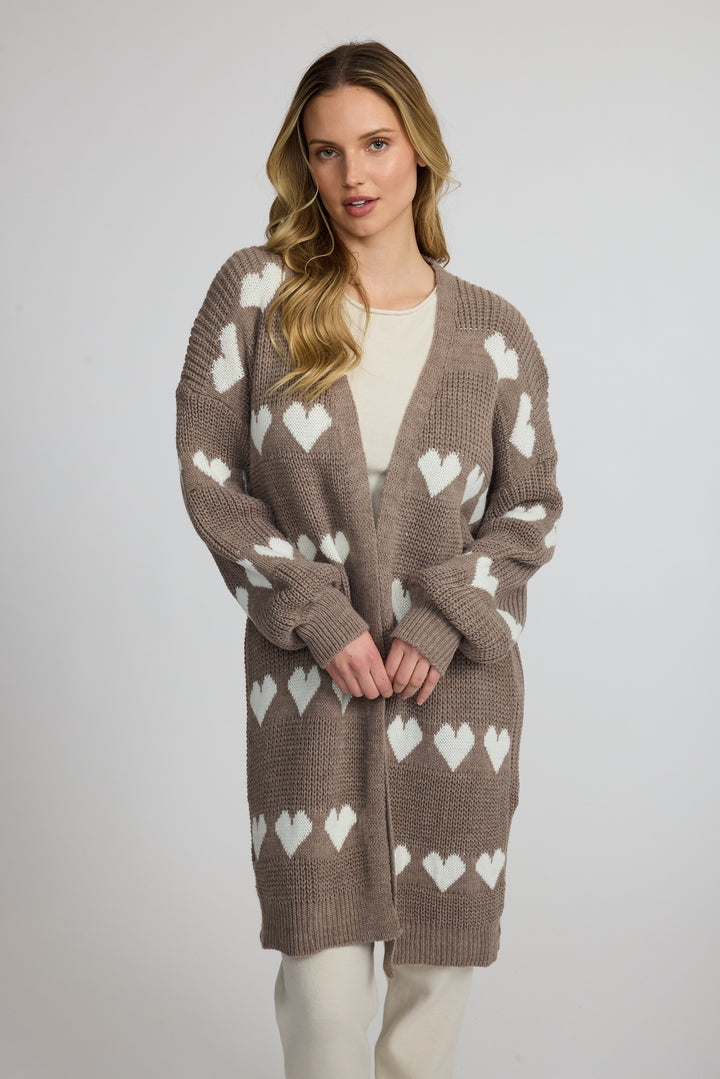 A woman modeling a brown open-front cardigan with a white heart pattern, worn over a cream top and cream pants against a light gray studio background.