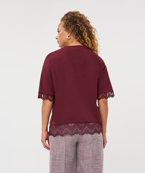 A woman viewed from the back wearing a burgundy short-sleeve top with lace trim and wide-leg mauve-gray trousers, standing against a plain light gray studio background.