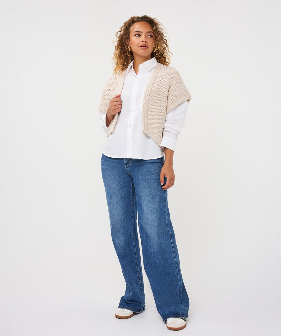 A woman modeling a white button-up shirt, a beige knit shrug, and wide-leg blue jeans against a plain white studio background.