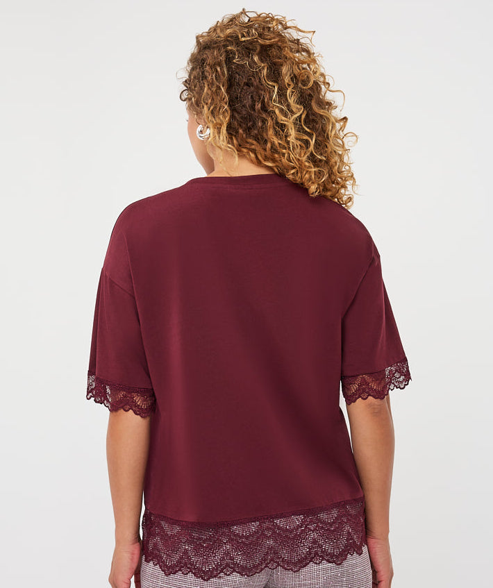 A woman viewed from the back wearing a burgundy short-sleeve top with lace trim at the cuffs and hem, paired with mauve-gray textured pants, against a plain light gray studio background.