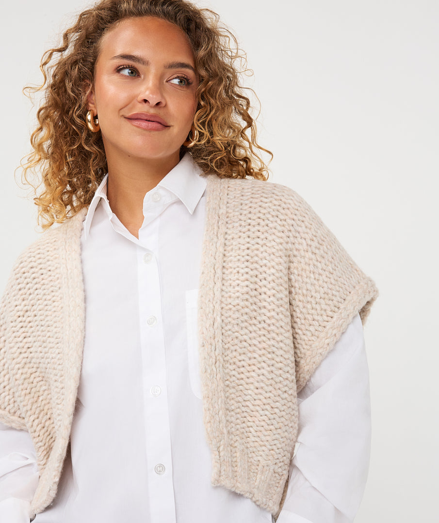A woman with curly blonde hair wearing a chunky knit beige shawl cardigan over a white button-down shirt, photographed against a clean white studio background.