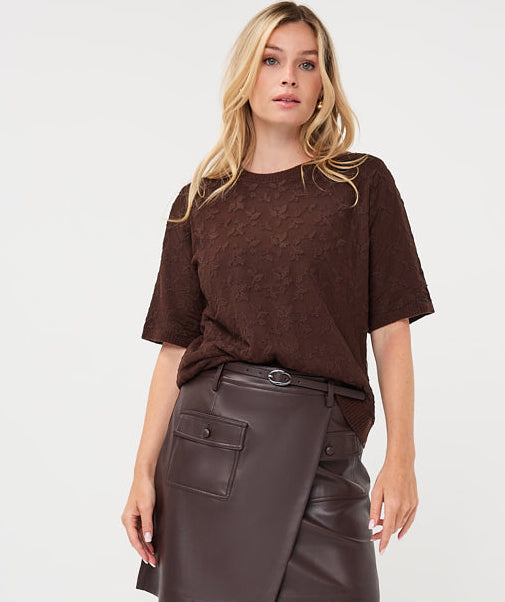 A woman modeling a dark brown short-sleeve knit top, a dark brown faux-leather wrap mini skirt, sheer black tights, and dark brown knee-high boots, against a plain white studio background.