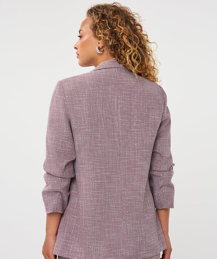 A woman with curly blonde hair is shown from the back wearing a textured mauve-purple blazer and matching trousers against a plain white studio background.
