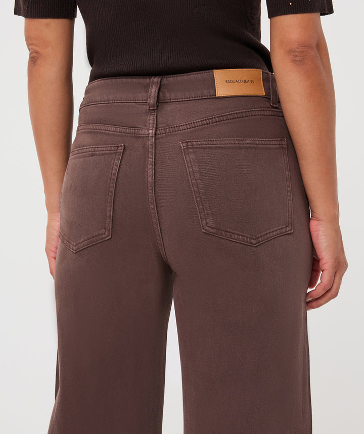 Rear view of a person wearing brown wide-leg jeans with a tan leather Kava Brand patch on the waistband, paired with a black ribbed top against a light gray studio background.