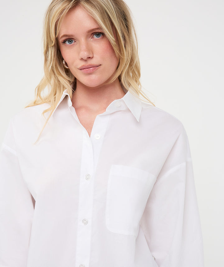 A blonde woman wearing a white long-sleeve button-up shirt with a chest pocket, photographed against a clean white studio background.