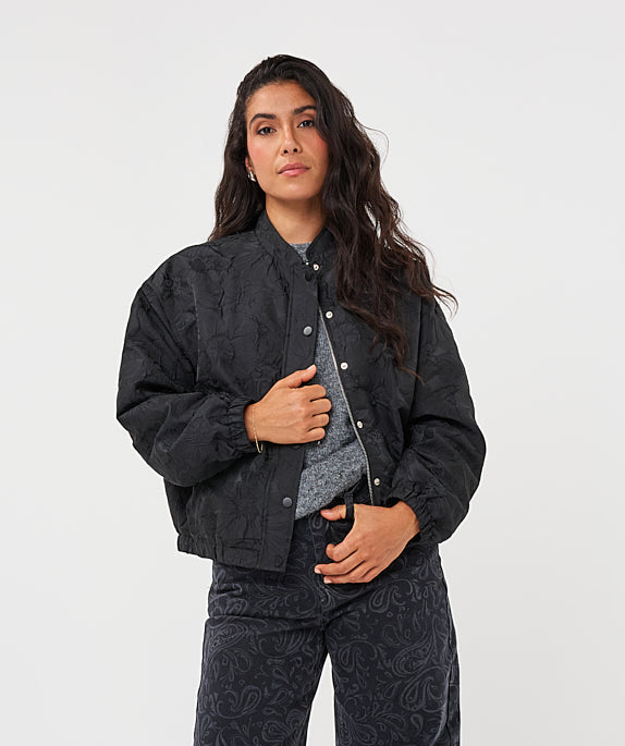 A woman modeling a black two-piece set featuring a cropped button-front jacket and matching wide-leg pants with a subtle paisley jacquard pattern, styled with a gray patterned shirt and black pointed-toe heels against a white studio background.
