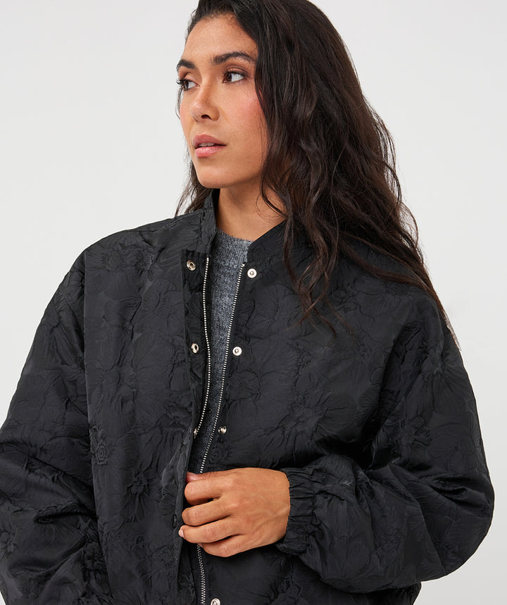 A woman wearing a black quilted bomber jacket with a textured, embossed pattern, shown in a close-up shot against a light gray background.