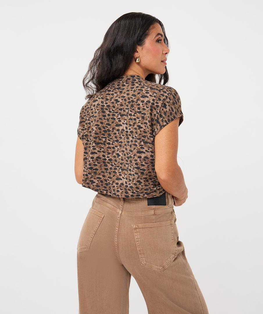 A woman shown from the back wearing a short-sleeve leopard-print crop top and tan pants against a plain light gray studio background.