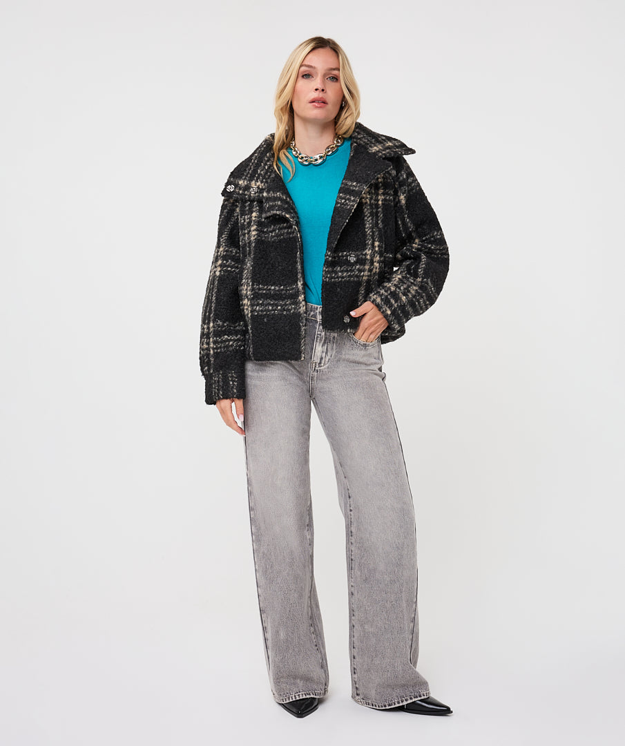 A model stands against a light gray studio background wearing a black and gray plaid cropped jacket over a bright turquoise top, paired with wide-leg light gray jeans and black pointed shoes.