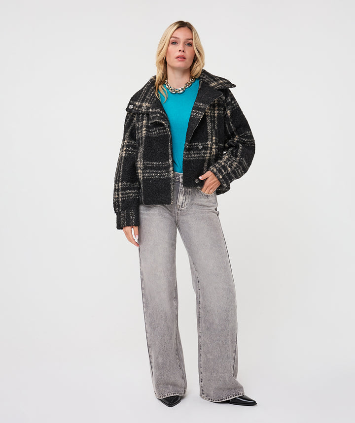 A model stands against a light gray studio background wearing a black and gray plaid cropped jacket over a bright turquoise top, paired with wide-leg light gray jeans and black pointed shoes.