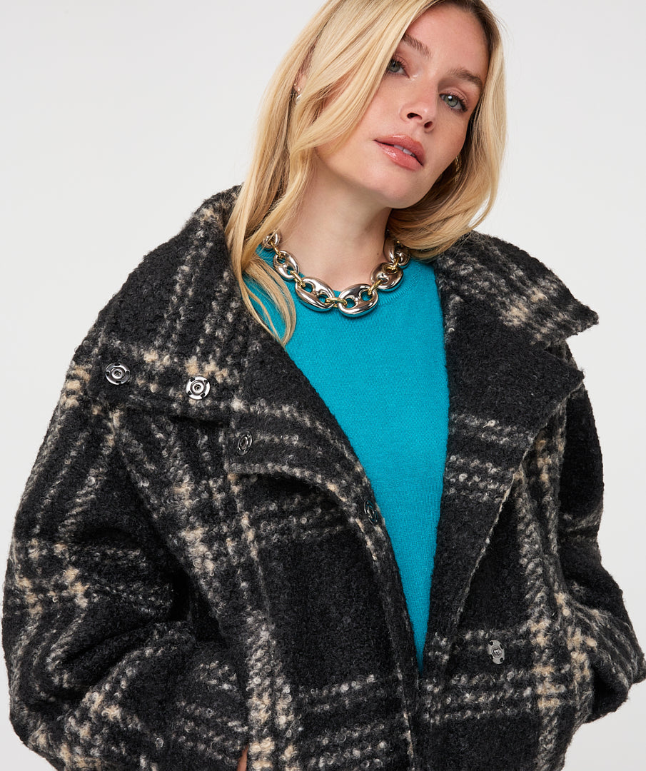 A close-up of a woman wearing a black and beige plaid textured coat over a bright turquoise top, accessorized with a chunky gold chain necklace, against a plain light gray background.