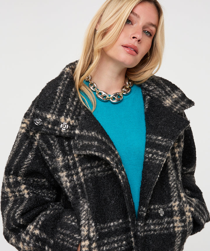 A close-up of a woman wearing a black and beige plaid textured coat over a bright turquoise top, accessorized with a chunky gold chain necklace, against a plain light gray background.