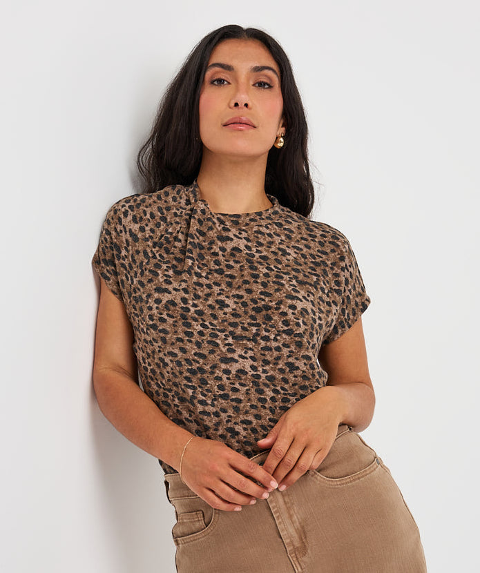 A woman wearing a short-sleeve leopard-print top and beige wide-leg jeans, posing against a plain white wall.