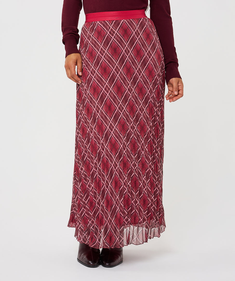 A woman wearing a long pleated maxi skirt with a red and white diamond pattern, paired with a matching red belt and dark burgundy top, shown against a plain white studio background.