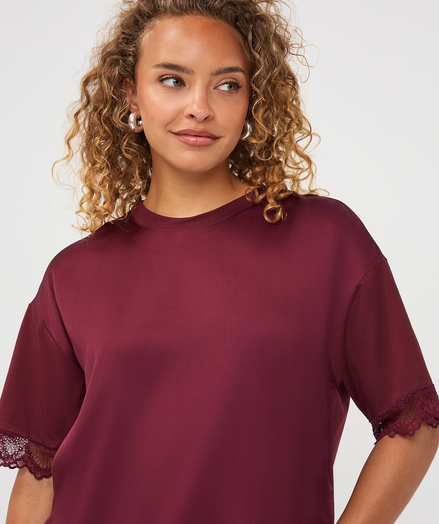 A woman with curly blonde hair wearing a short-sleeve crewneck top in deep burgundy with lace trim at the cuffs, photographed against a clean light gray studio background.
