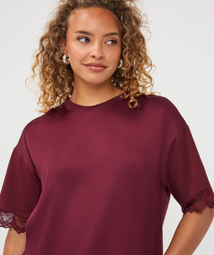 A woman with curly blonde hair wearing a short-sleeve crewneck top in deep burgundy with lace trim at the cuffs, photographed against a clean light gray studio background.
