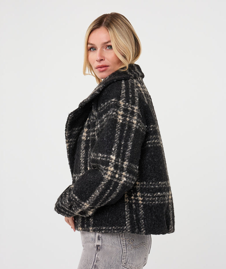 A woman wearing a dark charcoal and beige plaid cropped jacket, paired with light gray jeans, photographed against a plain light gray studio background.
