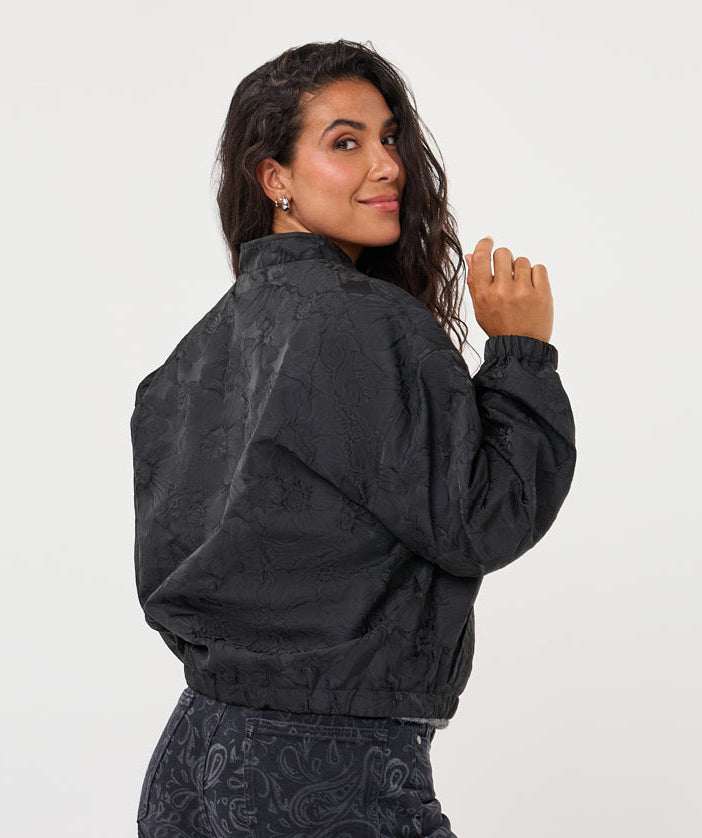 A woman wearing a black textured bomber jacket and matching black paisley-print pants, photographed from the back against a plain light gray studio background.