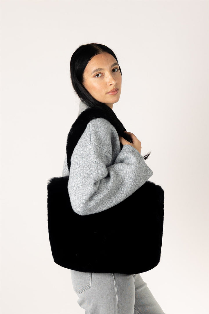 A woman wearing a gray sweater and light gray jeans carries a large black fuzzy shoulder bag against a plain white studio background.