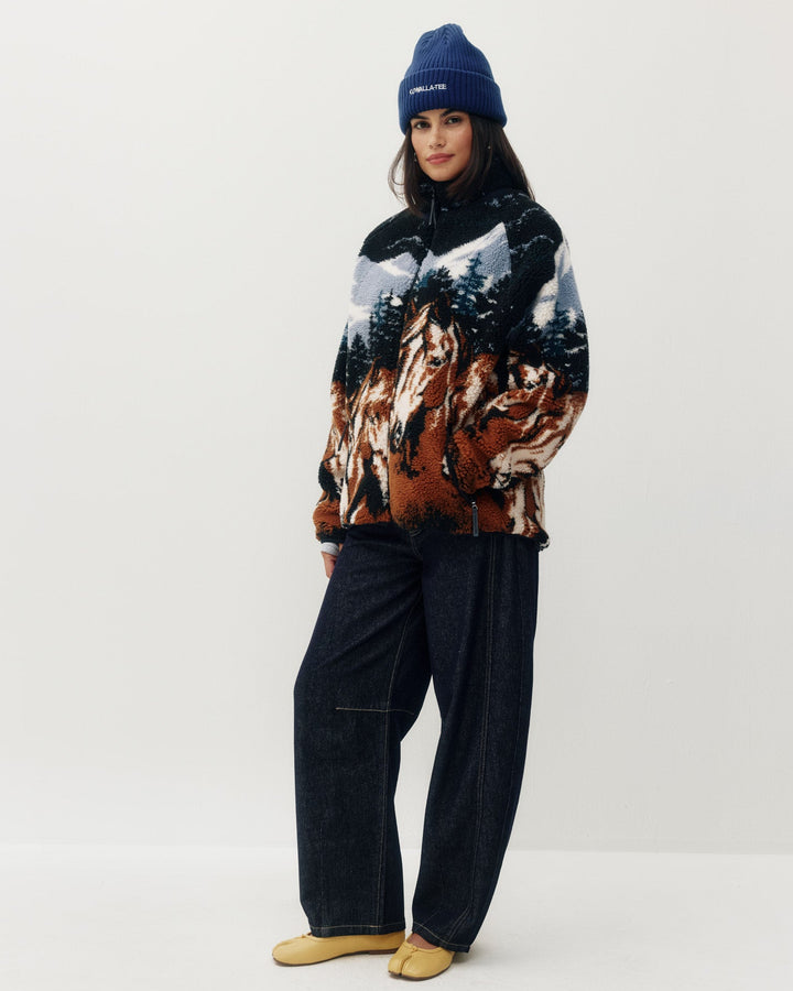 A model stands against a light gray studio background wearing a dark blue beanie with a white logo, a multicolor patterned fleece pullover featuring horses and trees, dark wide-leg jeans, and tan slip-on shoes.