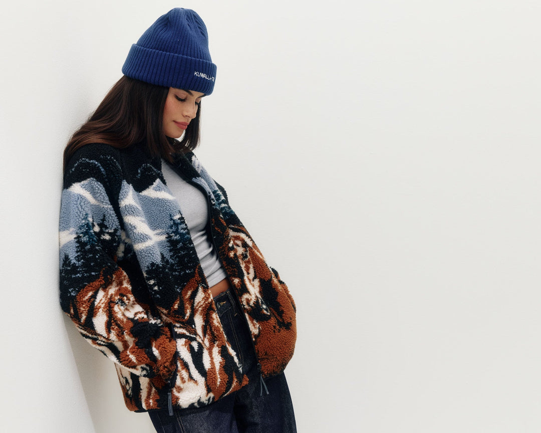 A woman wearing a blue knit beanie and a patterned knit jacket with a mountain and tree design, leaning against a plain white wall.