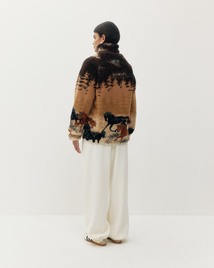 A model shown from the back wearing a brown and tan knit sweater with a black and brown horse pattern, paired with wide-leg cream trousers and a black beanie, against a plain white studio background.