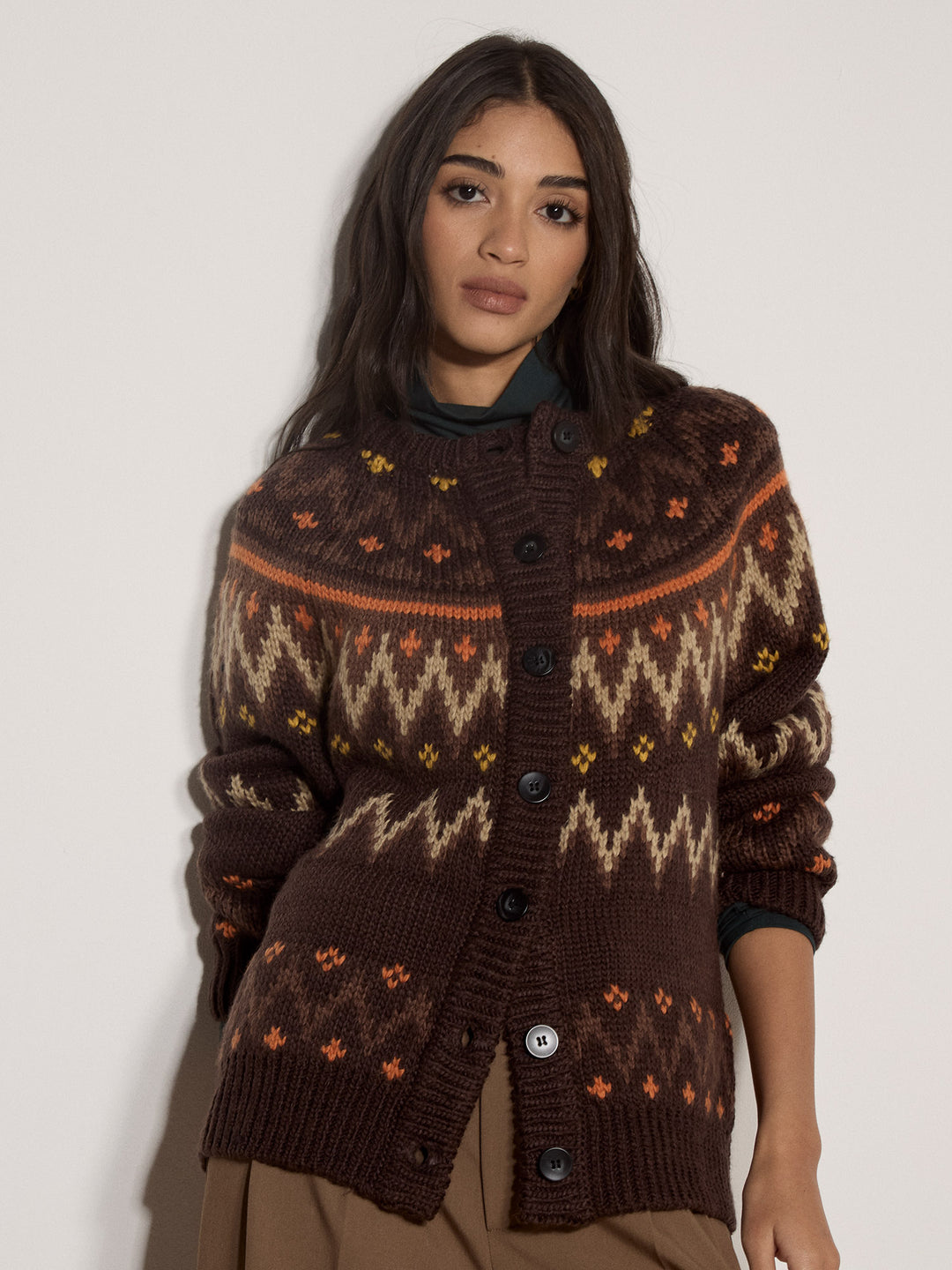 A woman wearing a brown knit button-up cardigan with Fair Isle patterns in beige, orange, and yellow, styled over a dark green turtleneck and brown trousers, against a plain light gray background.