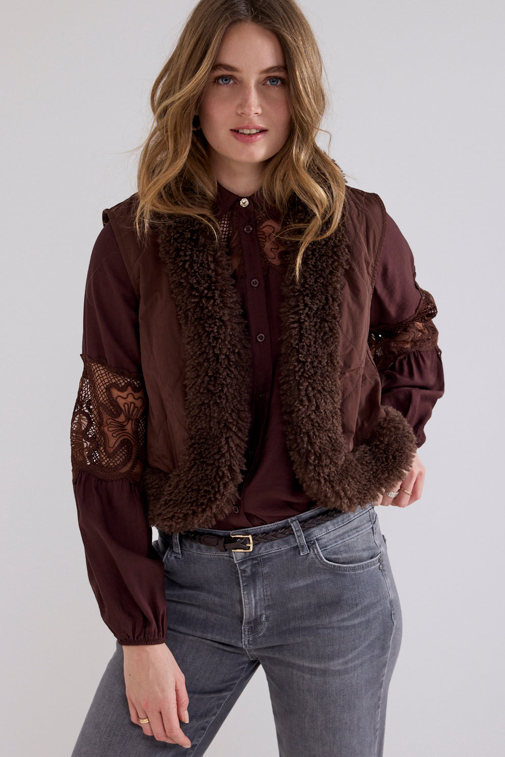A woman modeling a dark brown jacket with a shawl collar, lace sleeve panels, and a satin front, paired with gray jeans against a light gray studio background.