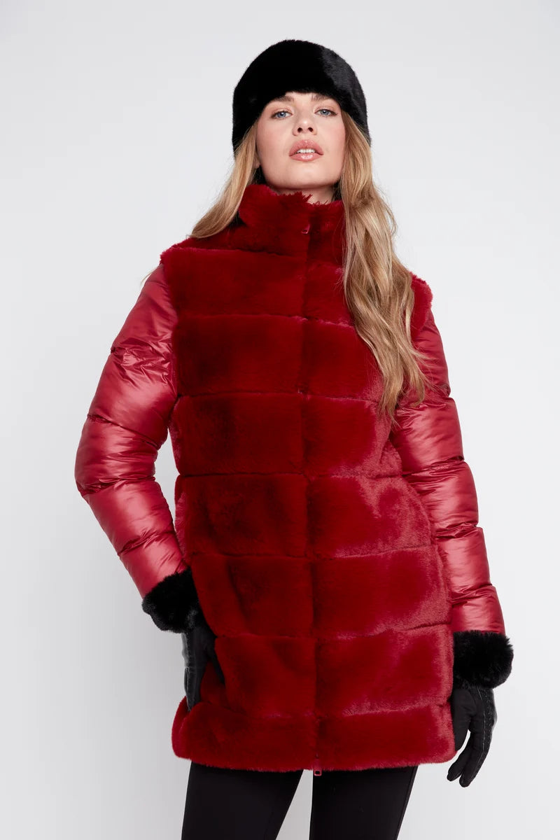 A woman modeling a long red faux fur coat with shiny red sleeves, a black beanie, and black gloves against a light gray studio background.