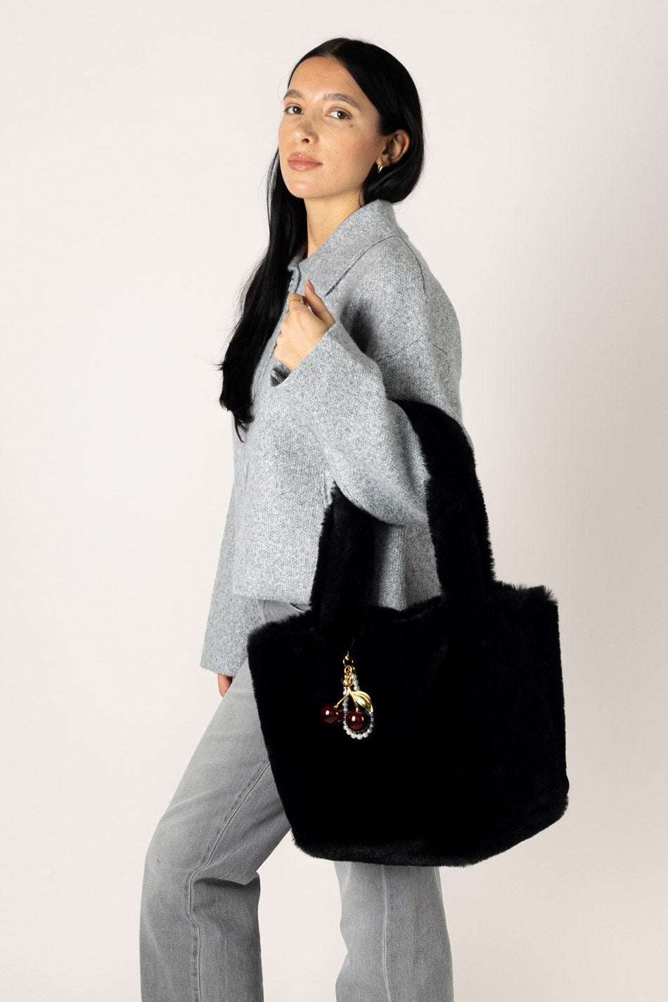A woman wearing a light gray knit sweater, wide-leg jeans, and dark flats carries a large black fuzzy tote bag with a small embroidered character patch on the front, posed against a plain light gray studio background.