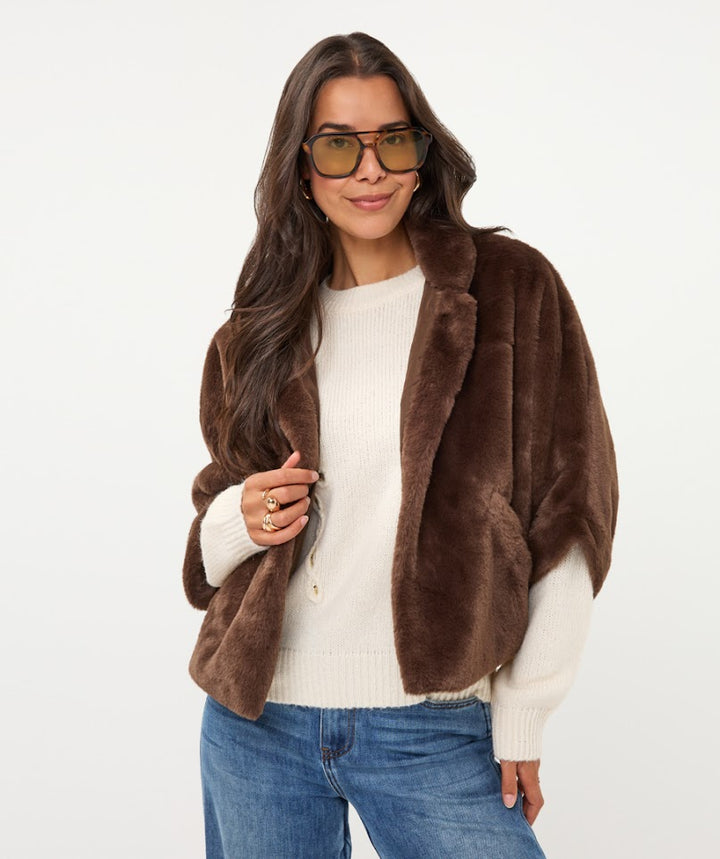 A woman modeling a brown faux fur jacket over a cream ribbed sweater and blue jeans, wearing tortoiseshell glasses, against a plain white background.