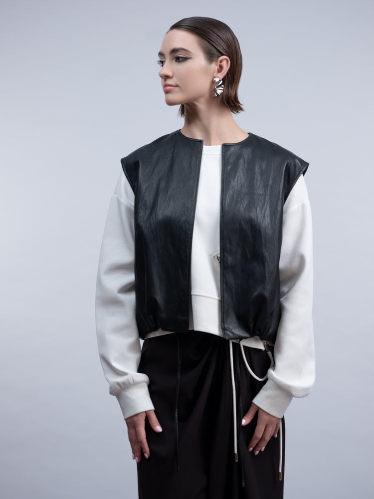 A model wearing a black faux-leather vest with white ribbed sleeves, layered over a white top and black drawstring pants, against a solid gray studio background.
