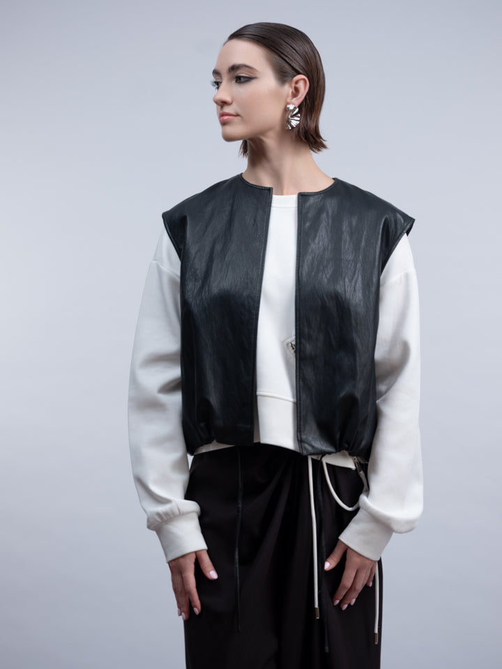 A model wearing a black faux-leather vest with white ribbed sleeves, layered over a white top and black drawstring pants, against a solid gray studio background.