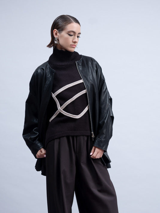 A model stands in a studio wearing a black turtleneck sweater with a white geometric stripe pattern, a black leather jacket, wide-leg black trousers, and black shoes.
