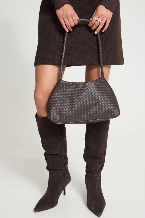 A model wearing a dark brown knit dress and matching over-the-knee boots holds a dark brown woven leather shoulder bag with a braided top handle against a light gray studio background.