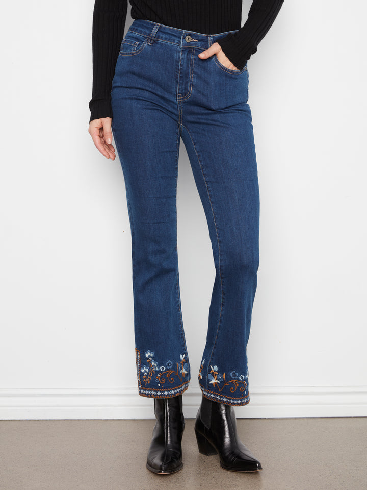 A person wearing dark blue denim jeans with floral embroidery near the hem, paired with a black long-sleeve top and black ankle boots, standing against a white wall.