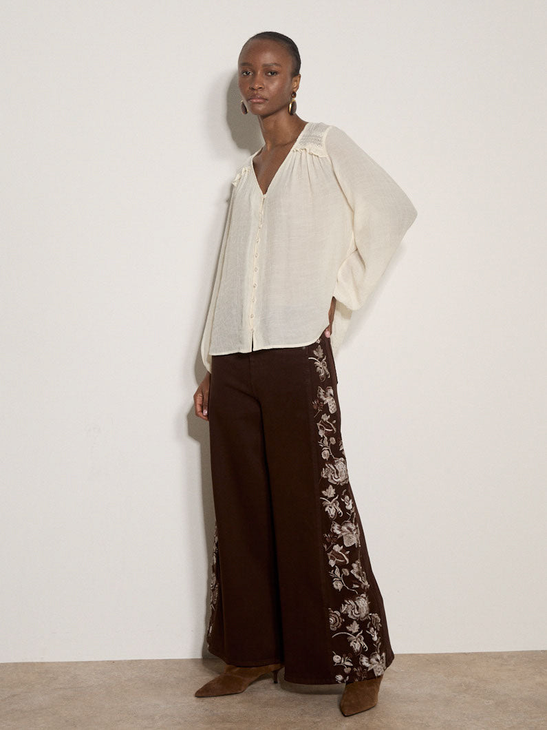 A model stands against a plain off-white wall wearing a loose off-white blouse with wide sleeves and dark brown wide-leg trousers featuring a white floral print down the sides, paired with brown pointed-toe flats.