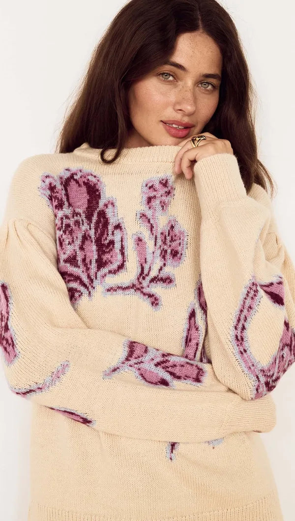 A woman wearing a cream-colored knit sweater with large purple and pink floral patterns, posed against a plain white background.