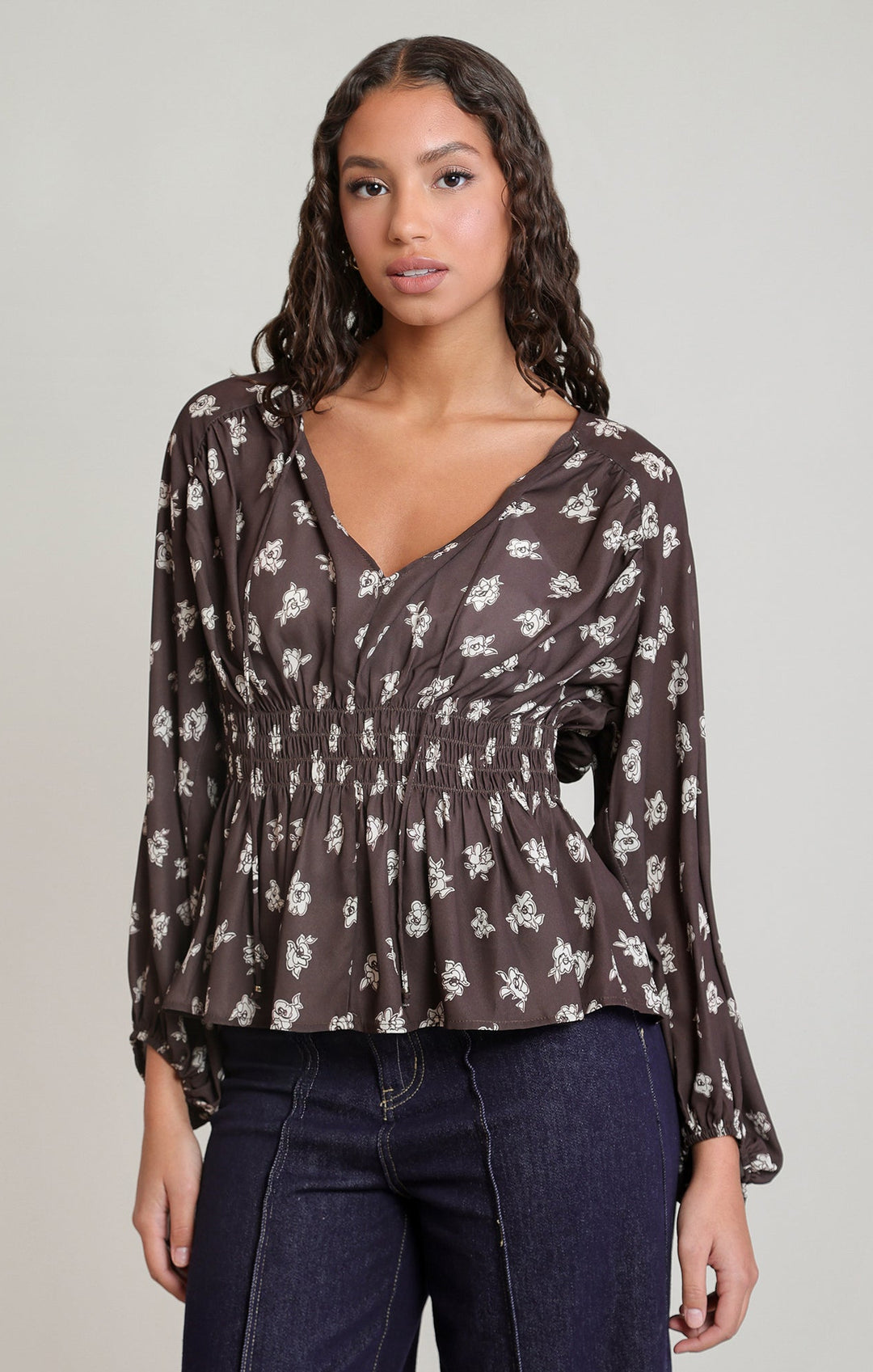 A woman modeling a dark brown long-sleeve blouse with a white floral pattern, paired with dark blue jeans, against a plain light gray studio background.