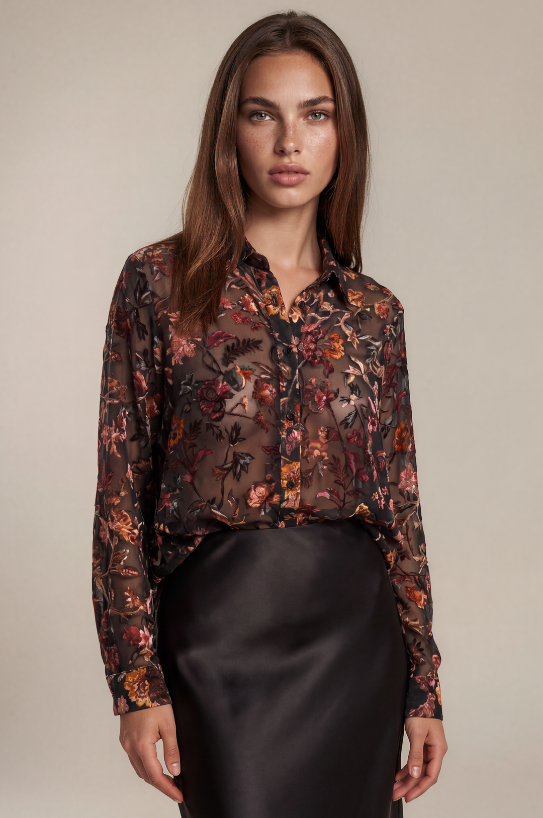 A woman modeling a sheer long-sleeve button-up blouse with an intricate floral and botanical print in dark brown, rust orange, and black, paired with a black satin skirt against a solid beige studio background.