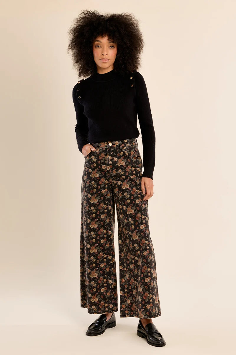 A model with voluminous dark curly hair stands against a warm beige studio background, wearing a black long-sleeve top with shoulder details and high-waisted, wide-leg floral pants in a brown, black, and beige pattern, paired with black leather loafers.