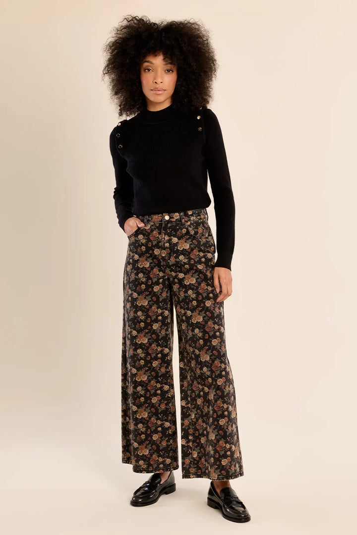 A model with voluminous dark curly hair stands against a warm beige studio background, wearing a black long-sleeve top with shoulder details and high-waisted, wide-leg floral pants in a brown, black, and beige pattern, paired with black leather loafers.